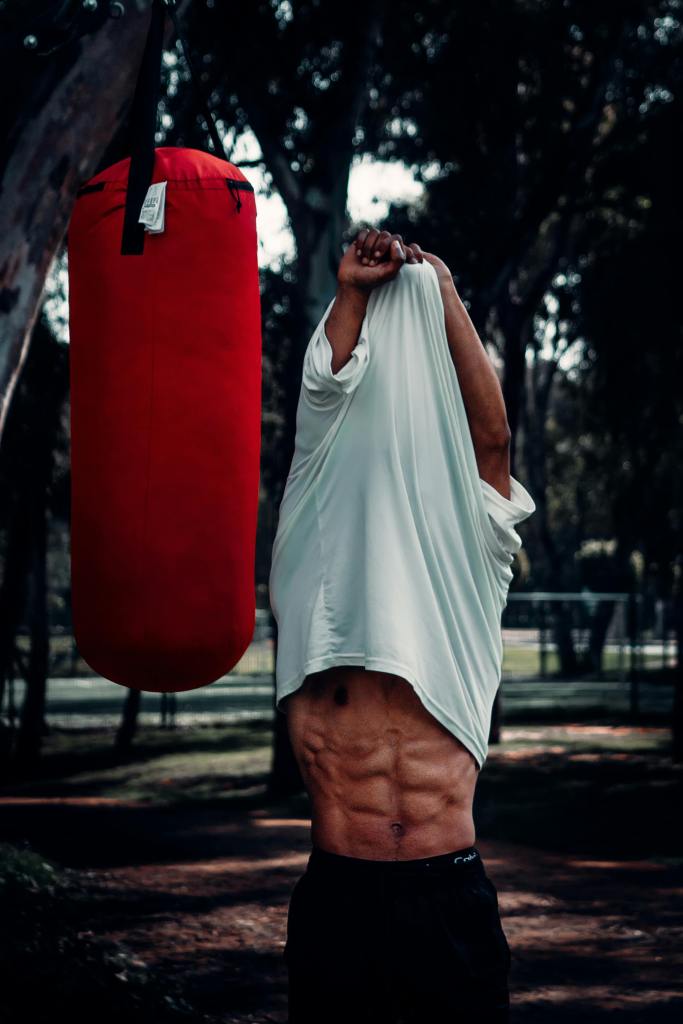A man takes off his shirt revealing a six pack while standing next to a punching bag.